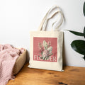 Tote bag with floral design and 'FLOWER' text on a wooden surface with a white wall background
