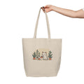 Beige tote bag with a cat and plant design held by a hand on a white background