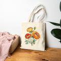 Tote bag with floral design on a wooden surface next to a basket and pink blanket.