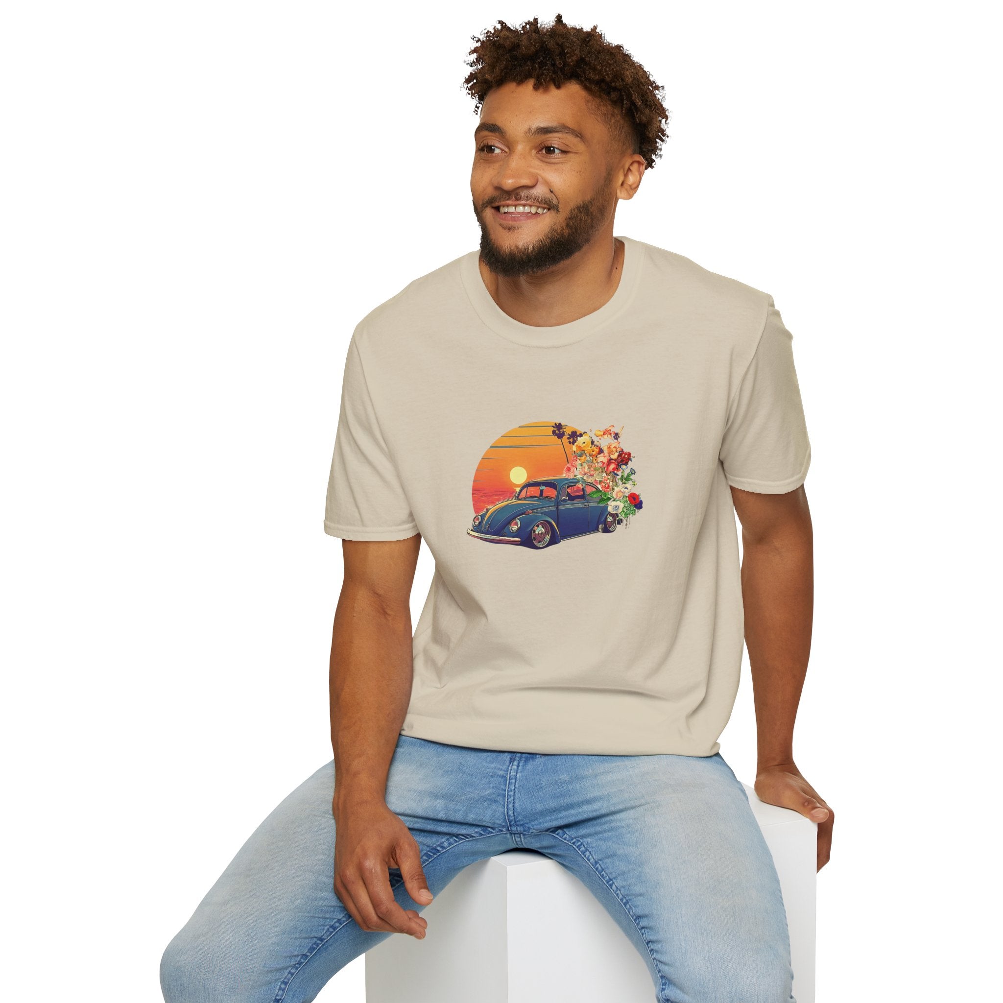 Man wearing a beige t-shirt with a graphic design of a vintage vw car and sunset, sitting on a white stool against a white background.