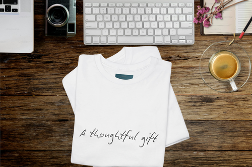 White t-shirt with 'A thoughtful gift' text on a wooden desk with laptop, keyboard, and coffee.