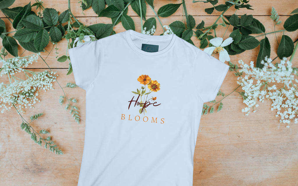 White t-shirt with floral design and 'Hope Blooms' text on a wooden surface with greenery and flowers.