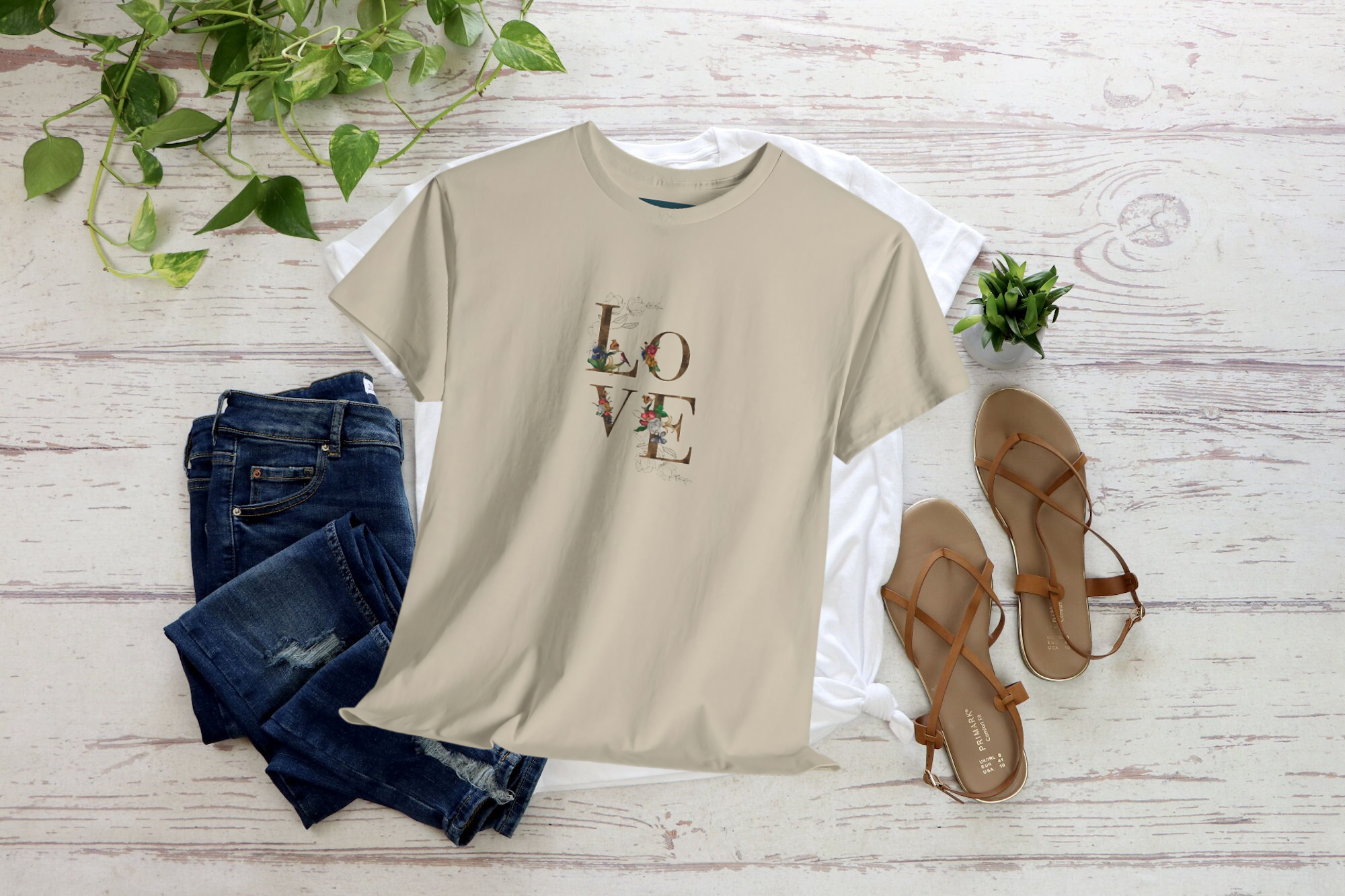 Beige sand t-shirt with 'LOVE' print, blue jeans, and brown sandals on a light wooden surface.