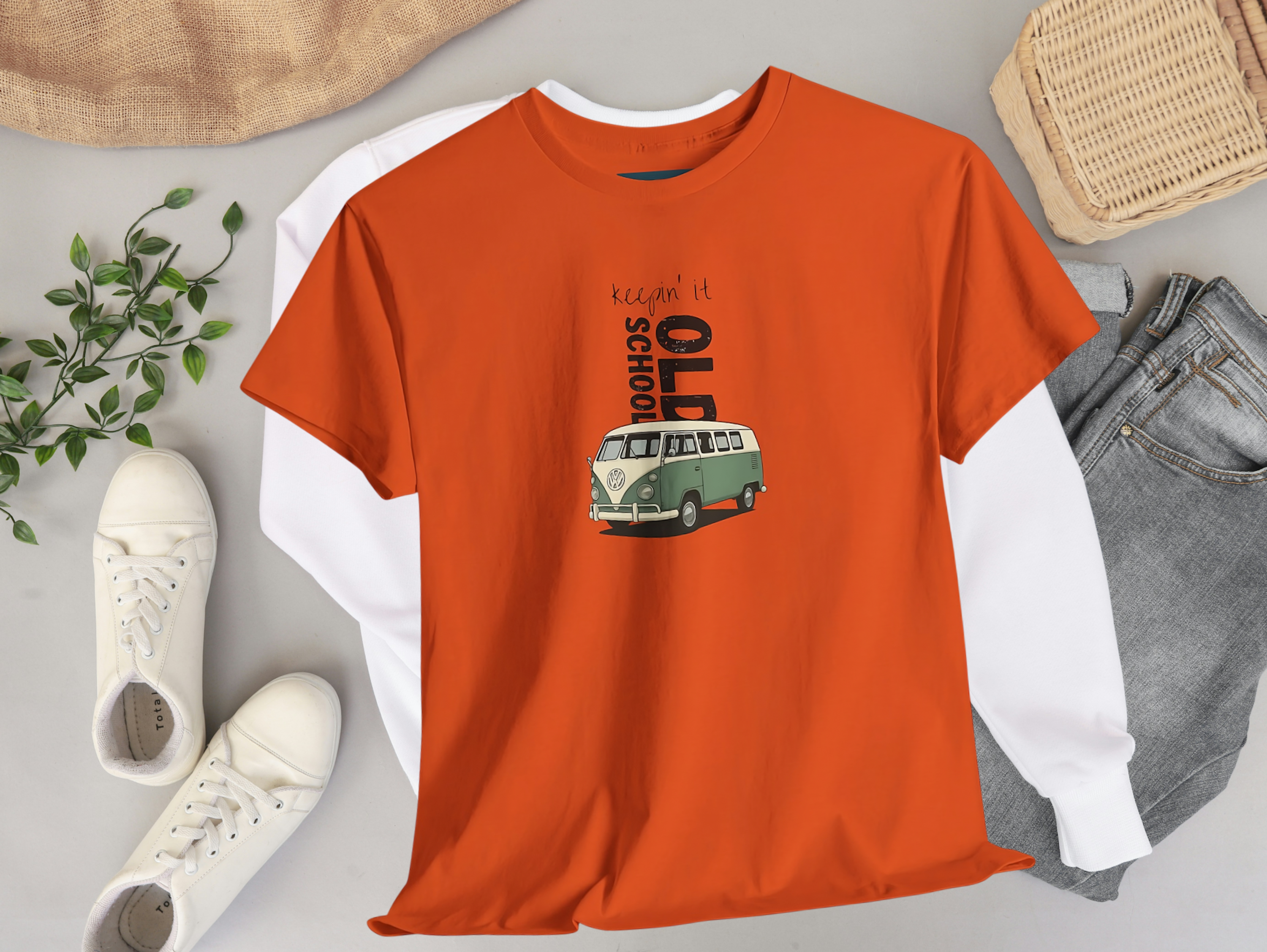 Orange t-shirt with VW bus graphic design on a gray surface with shoes, jeans, and a plant.