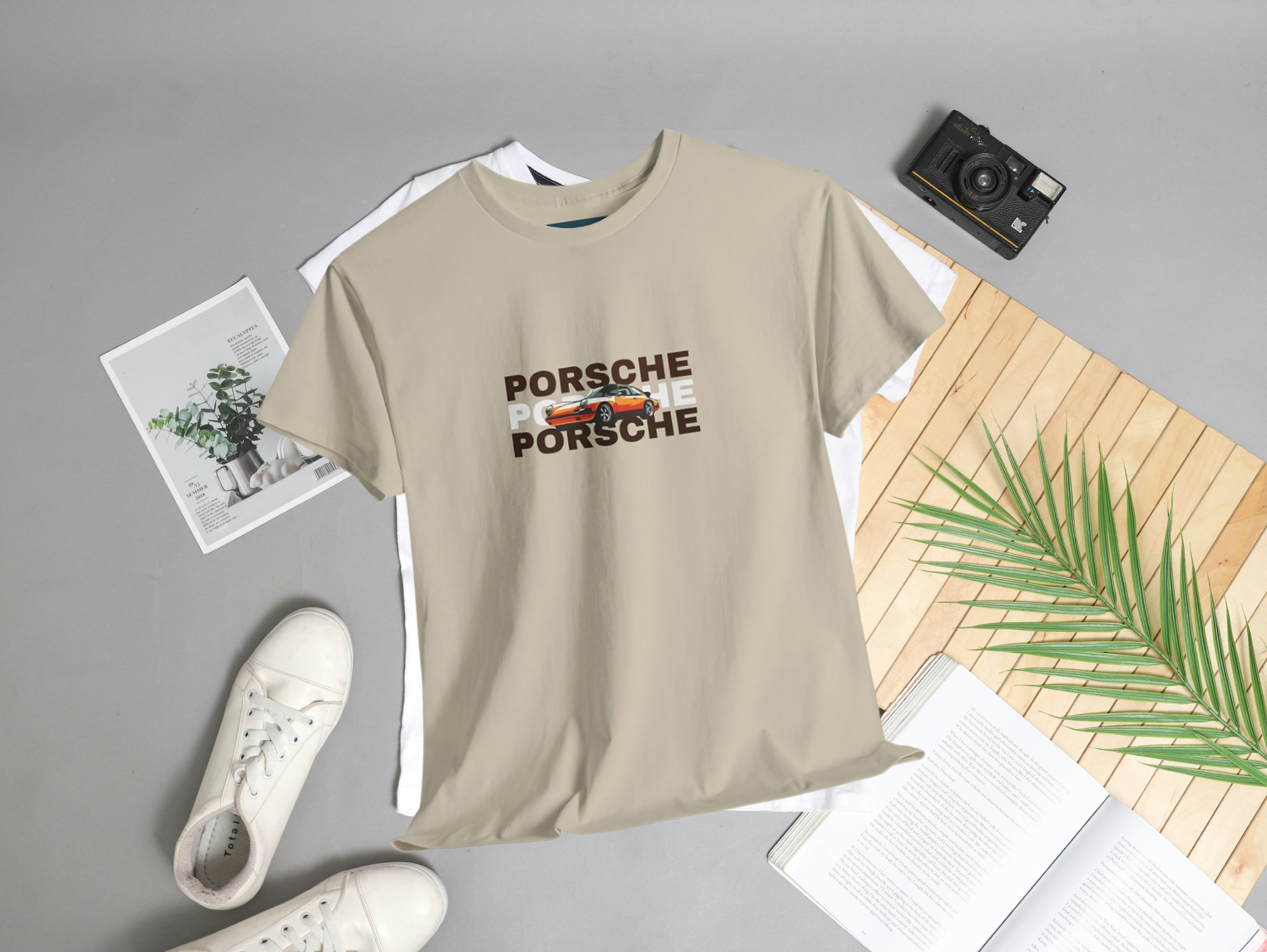Beige t-shirt with Porsche logo graphic on a gray surface with sneakers and a camera.