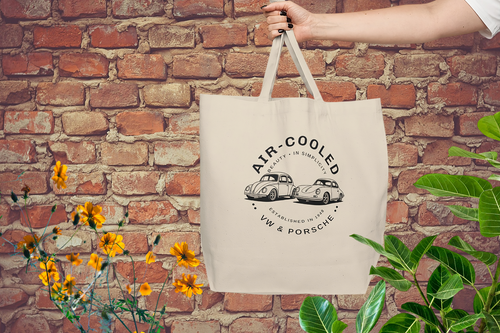 Beige tote bag with VW and Porsche design held against a brick wall with plants and flowers.