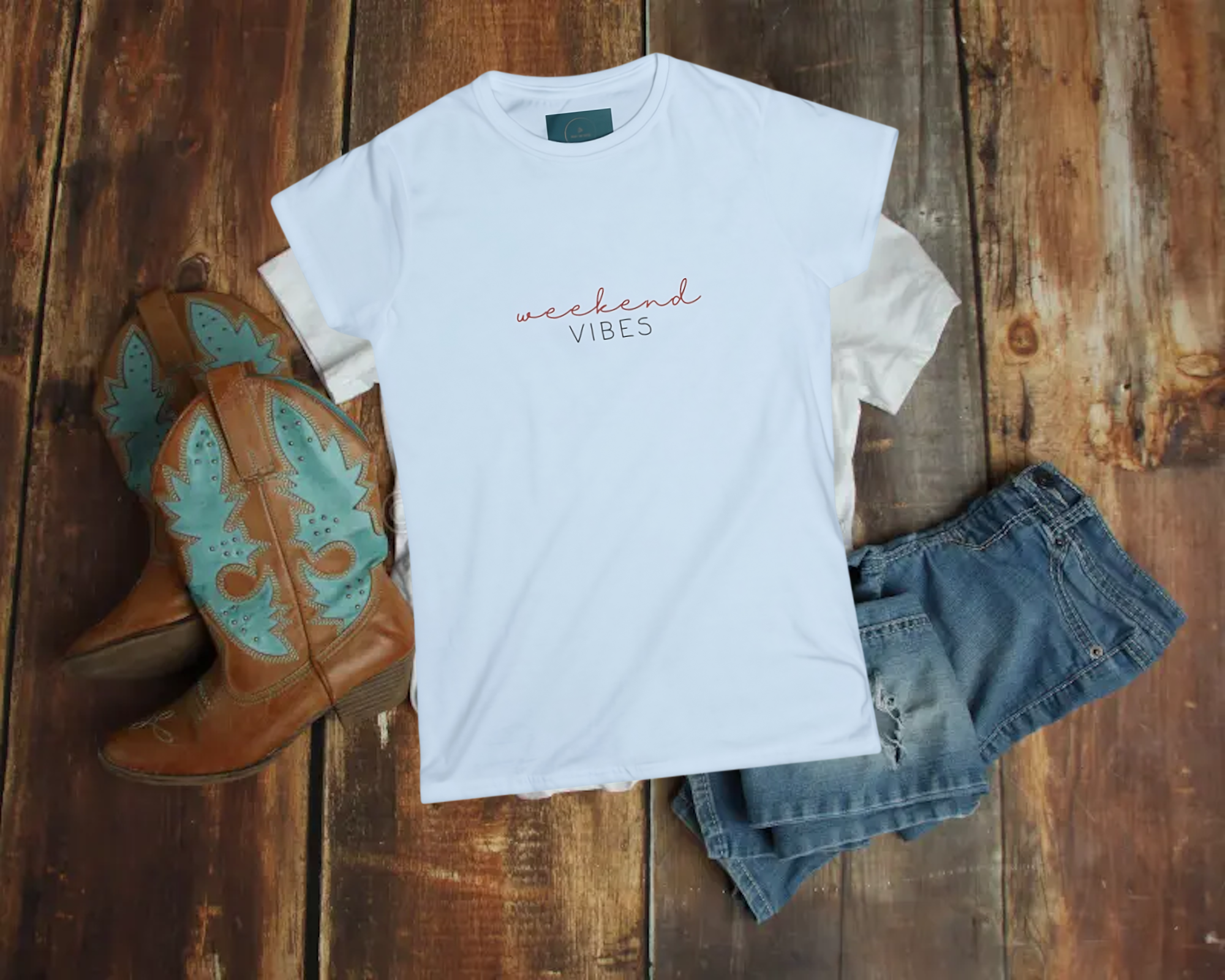 White t-shirt with 'weekend vibes' text, blue jeans, and brown boots on a wooden surface